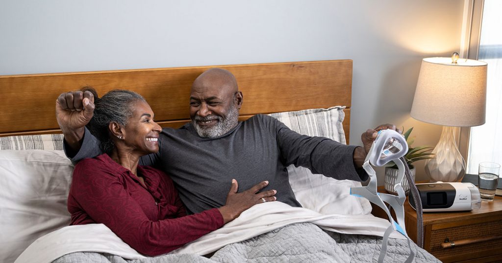 Couple smiling in bed where husband is getting used to using his new CPAP machine