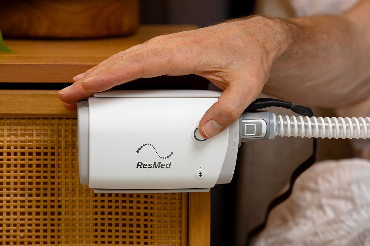 ResMed AirMini next to bed on nightstand clipped with hand pushing power button.