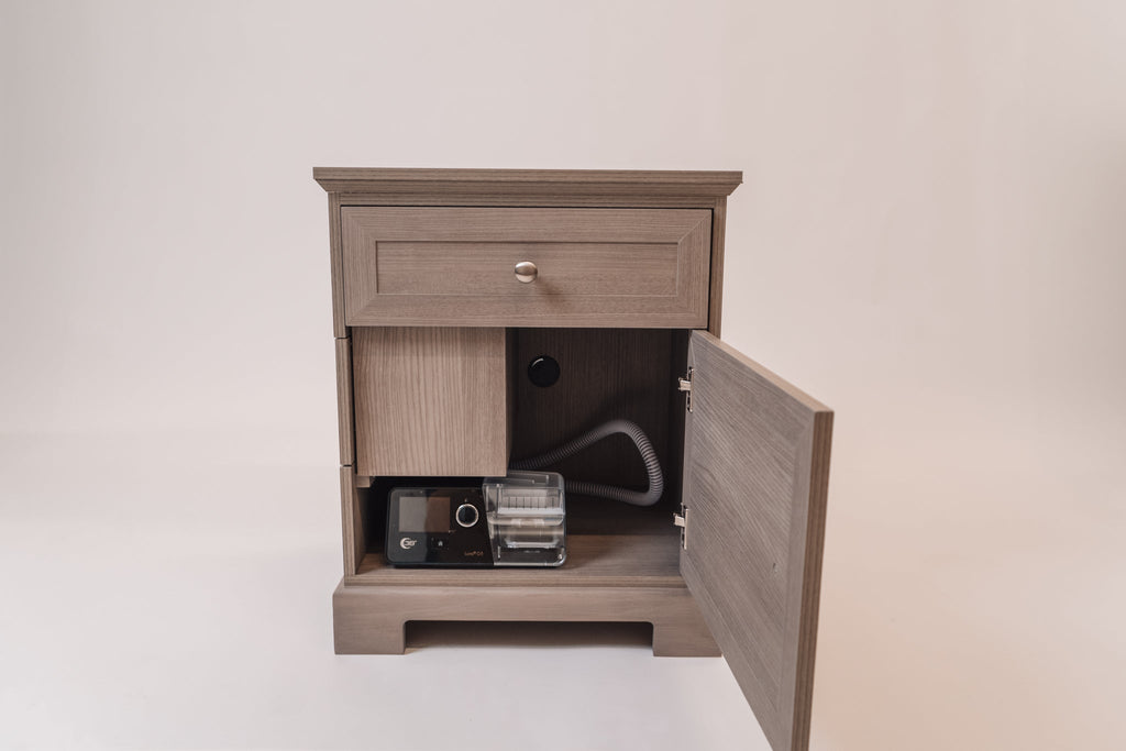 A front view of the Charleston Gray CPAP nightstand with the lower cabinet door open, showcasing an organized interior with a CPAP machine, hose storage, and a back panel cutout for cable management. The top drawer remains closed, maintaining the sleek aesthetic.
