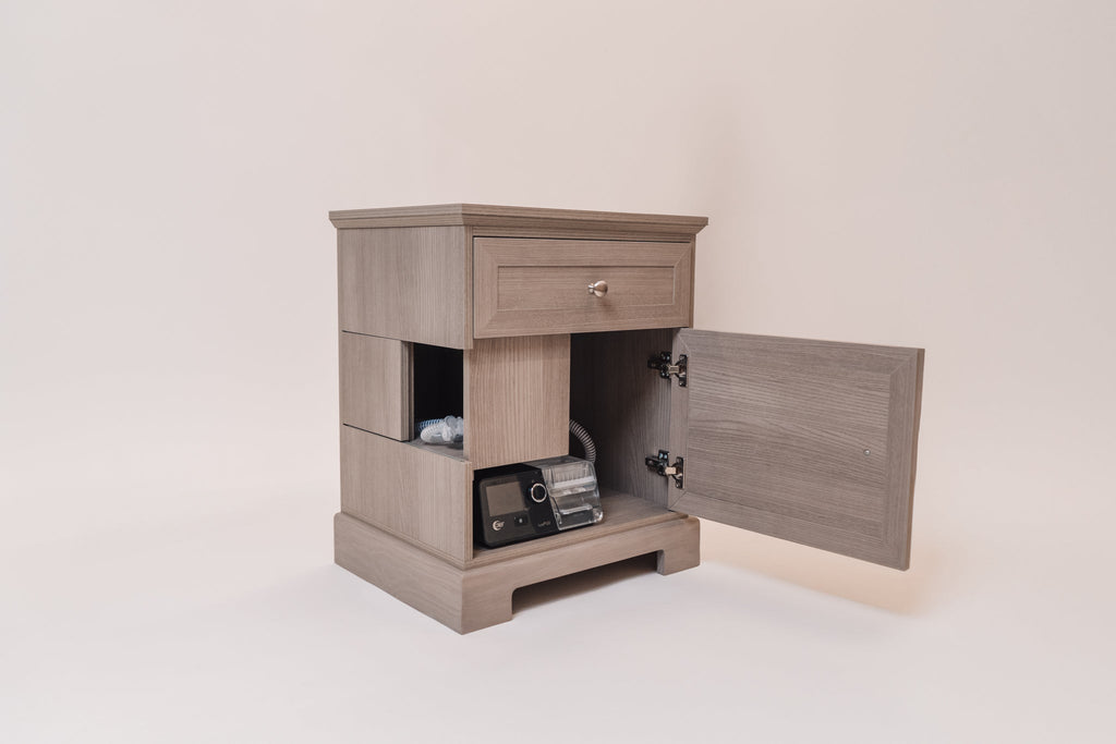 A Charleston Gray CPAP nightstand with its lower cabinet door open, revealing a black AirSense 10 CPAP machine and coiled hose neatly stored inside. The side cutouts allow for hose access, while the top drawer provides additional storage.