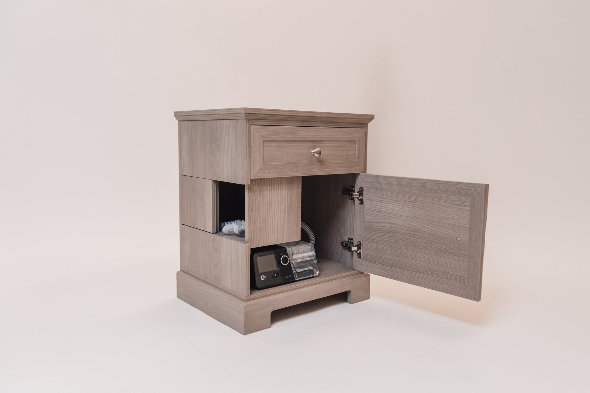 A Charleston Gray CPAP nightstand with its lower cabinet door open, revealing a black AirSense 10 CPAP machine and coiled hose neatly stored inside. The side cutouts allow for hose access, while the top drawer provides additional storage.