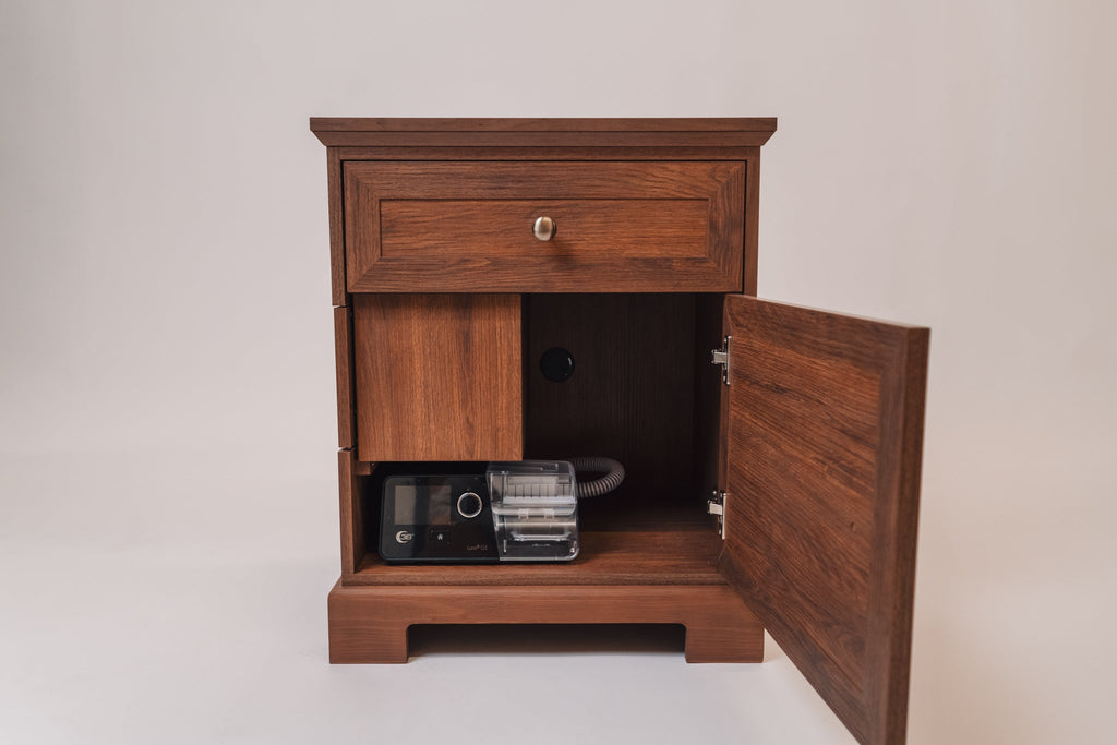 A CPAP nightstand with a warm walnut finish, shown with the lower cabinet door open. Inside, a black CPAP machine and coiled hose are neatly stored. The back panel has a circular cutout for cable management, and the nightstand features a traditional design with elegant molding details.