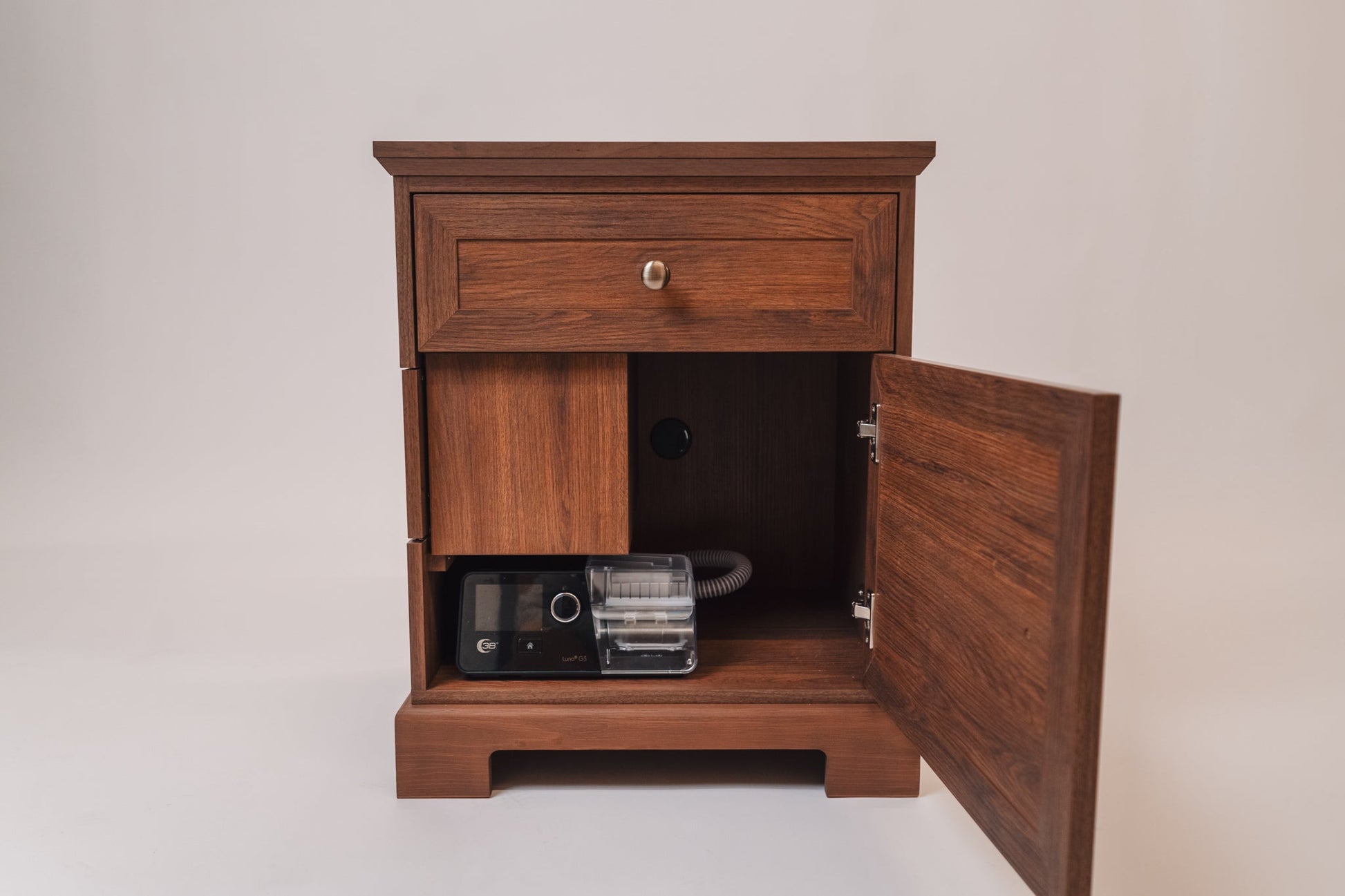 A CPAP nightstand with a warm walnut finish, shown with the lower cabinet door open. Inside, a black CPAP machine and coiled hose are neatly stored. The back panel has a circular cutout for cable management, and the nightstand features a traditional design with elegant molding details.