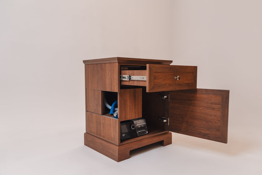 A CPAP nightstand in Charleston Walnut finish with a top drawer and a lower cabinet door open, revealing a CPAP machine and tubing storage. The nightstand features side cutouts for hose management, a pull-out drawer with metal slides, and a classic wood grain finish.