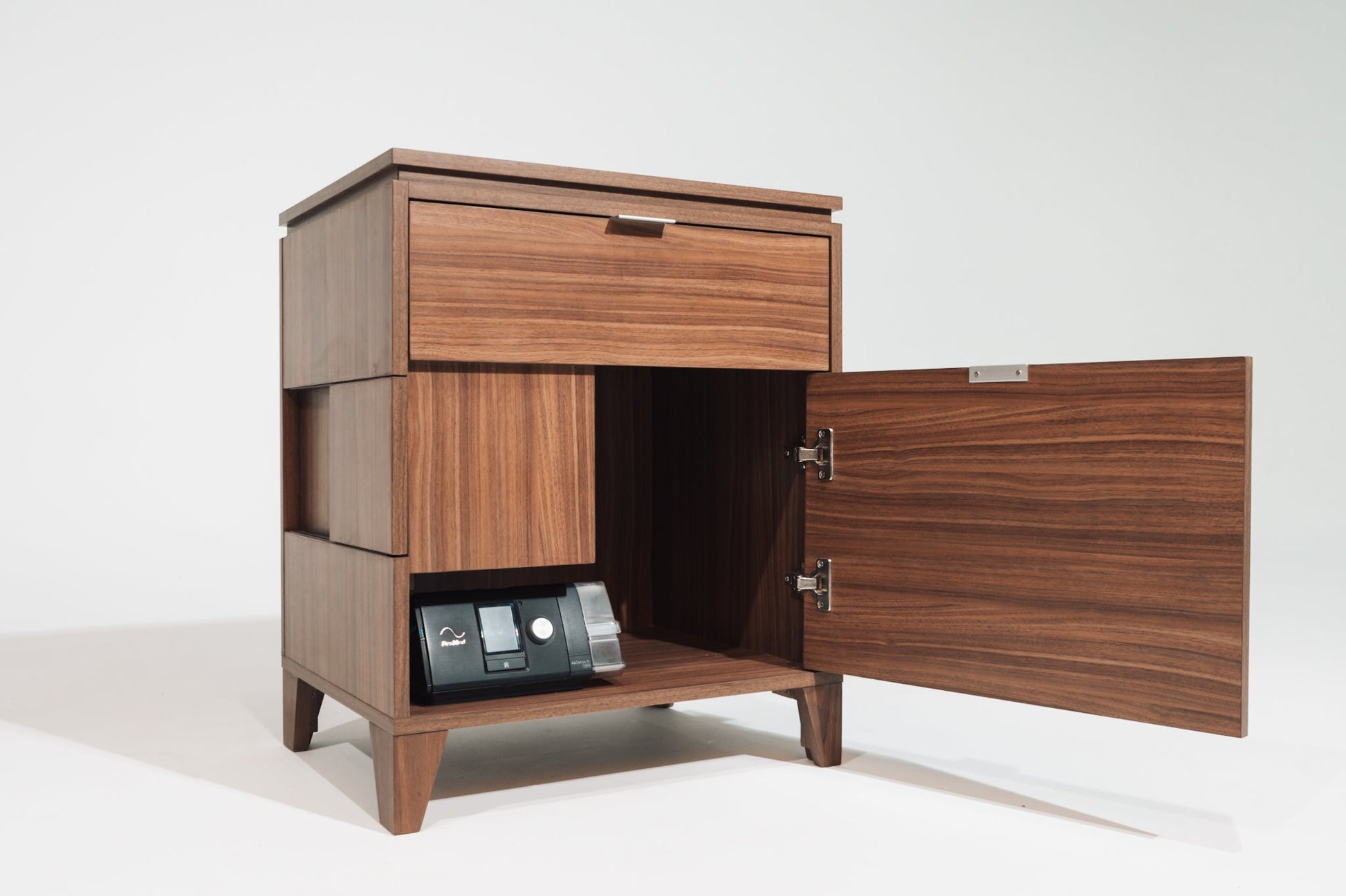 A CPAP nightstand in Soho Walnut with the lower cabinet door open, showcasing a black CPAP machine stored inside. The nightstand features a dedicated compartment for equipment storage, with a back panel cutout for cable management.