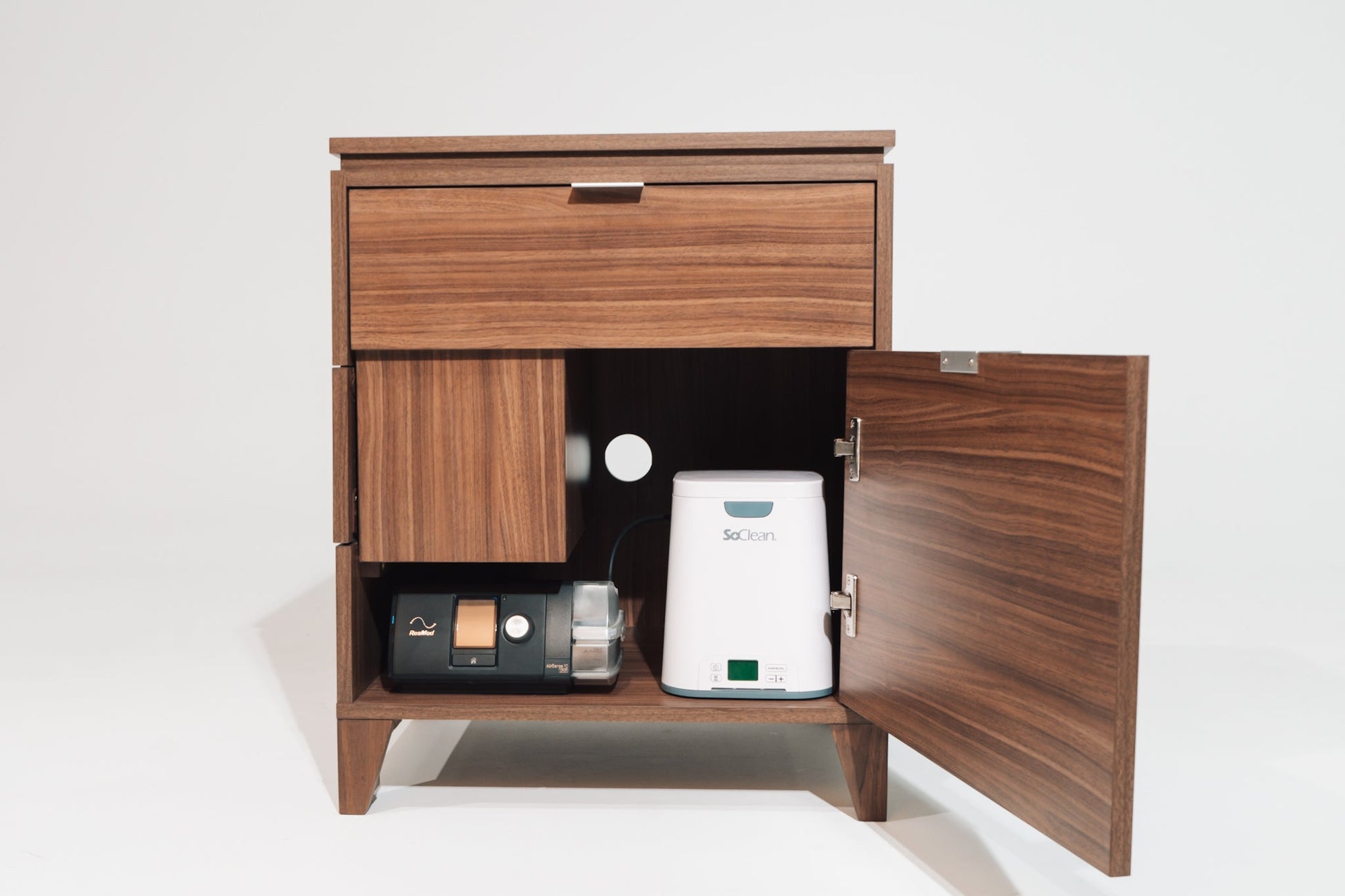 A front-facing view of the Soho Walnut CPAP nightstand with the lower cabinet door open, revealing a neatly stored CPAP machine and a white SoClean CPAP sanitizer. The interior is designed for organized storage, with proper ventilation and wire access.