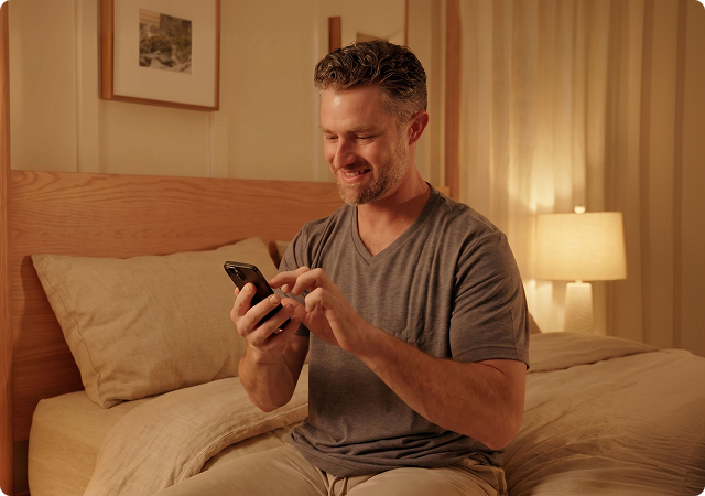 Man sitting in bed using a smartphone at night with a bedside lamp glowing in the background.