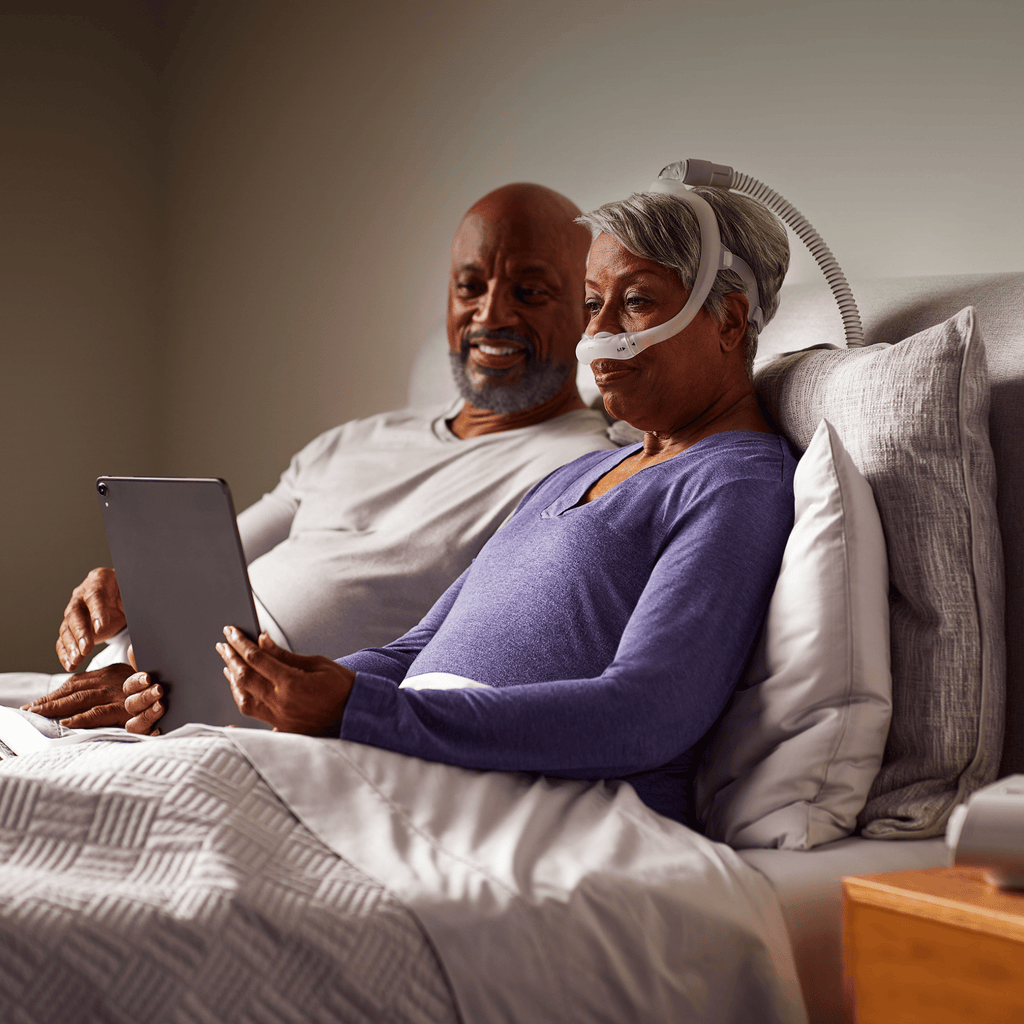Woman wearing philips dreamwear nasal pillow mask sits in bed with her partner as they review her dreammapper app scores on a tablet.