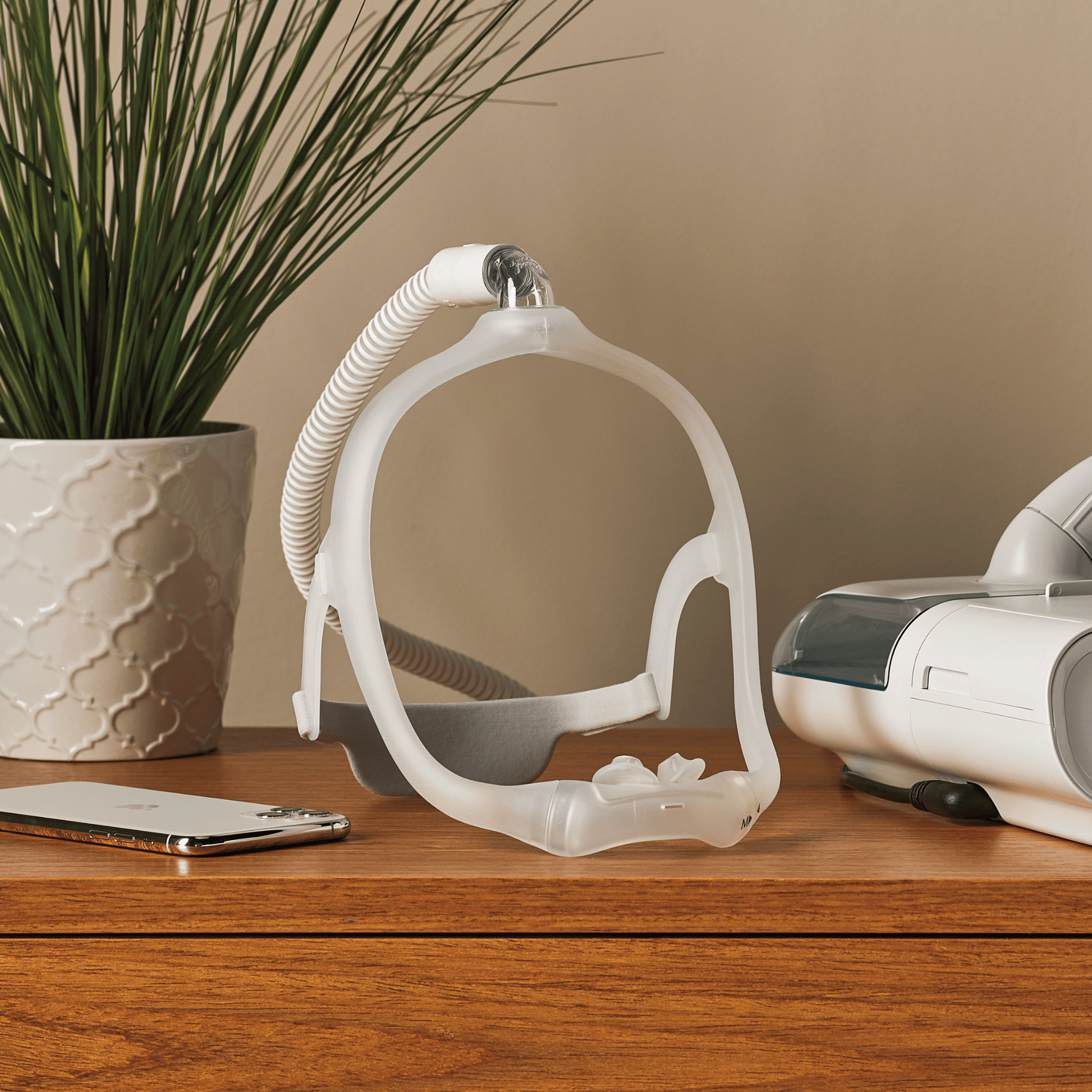 A dreamwear nasal pillow CPAP mask rests on a nightstand in soft lighting next to a Philips DreamStation CPAP machine, a smartphone, and a potted plant.