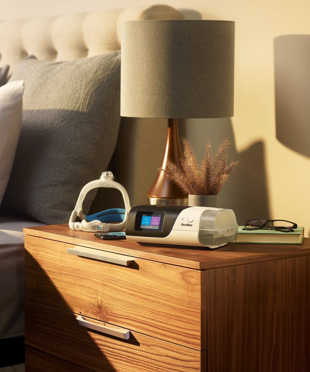 ResMed CPAP machine and mask on a wooden bedside table next to a lamp.