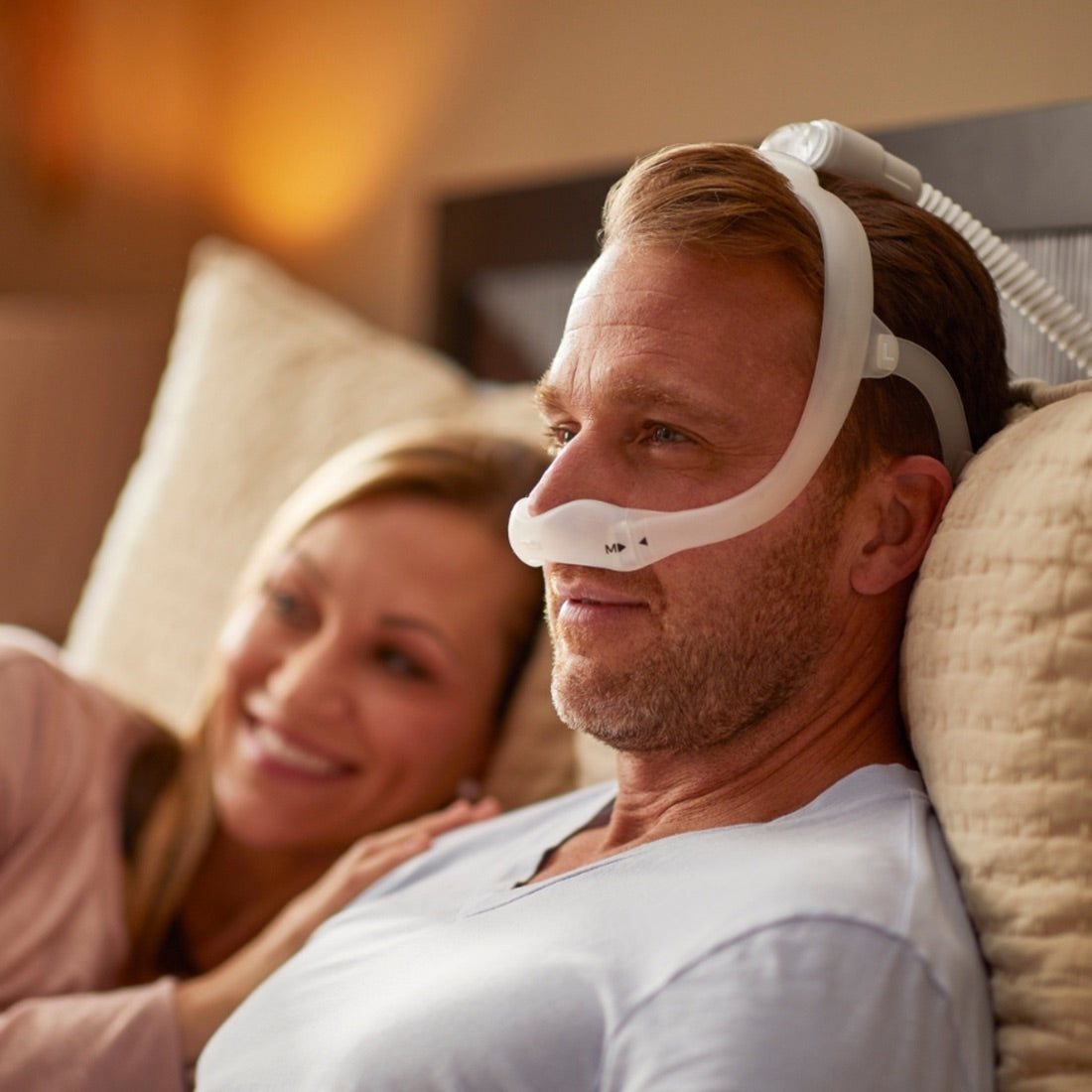 A man wearing a white CPAP nasal mask while lying in bed next to a smiling woman.
