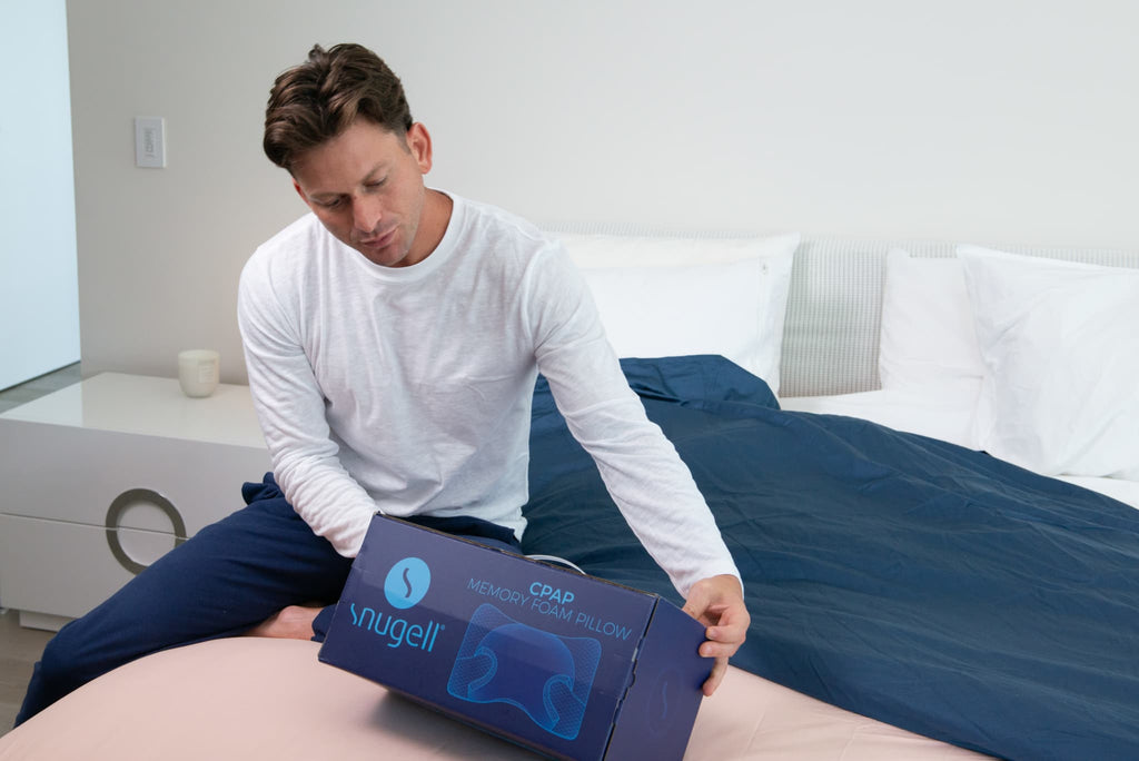 Man holding a Snugell CPAP Memory Foam Pillow box, sitting on a bed ready to unpack the sleep aid.