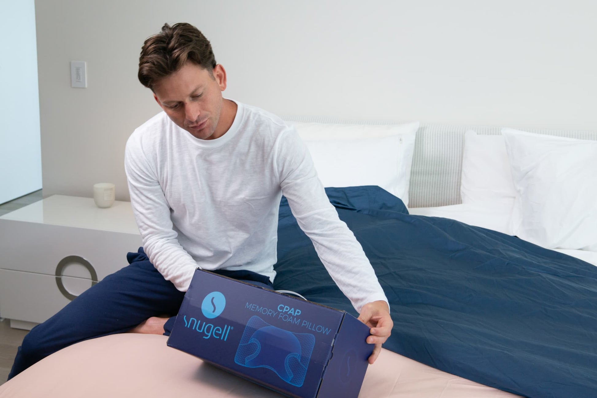 Man holding a Snugell CPAP Memory Foam Pillow box, sitting on a bed ready to unpack the sleep aid.