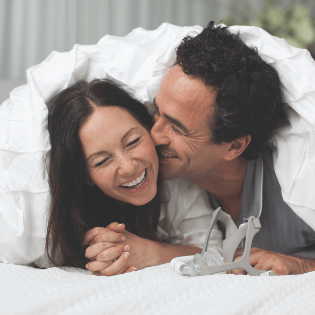 a couple laughs in bed under the covers with a wisp fabric nasal mask sitting on the bed in front of them