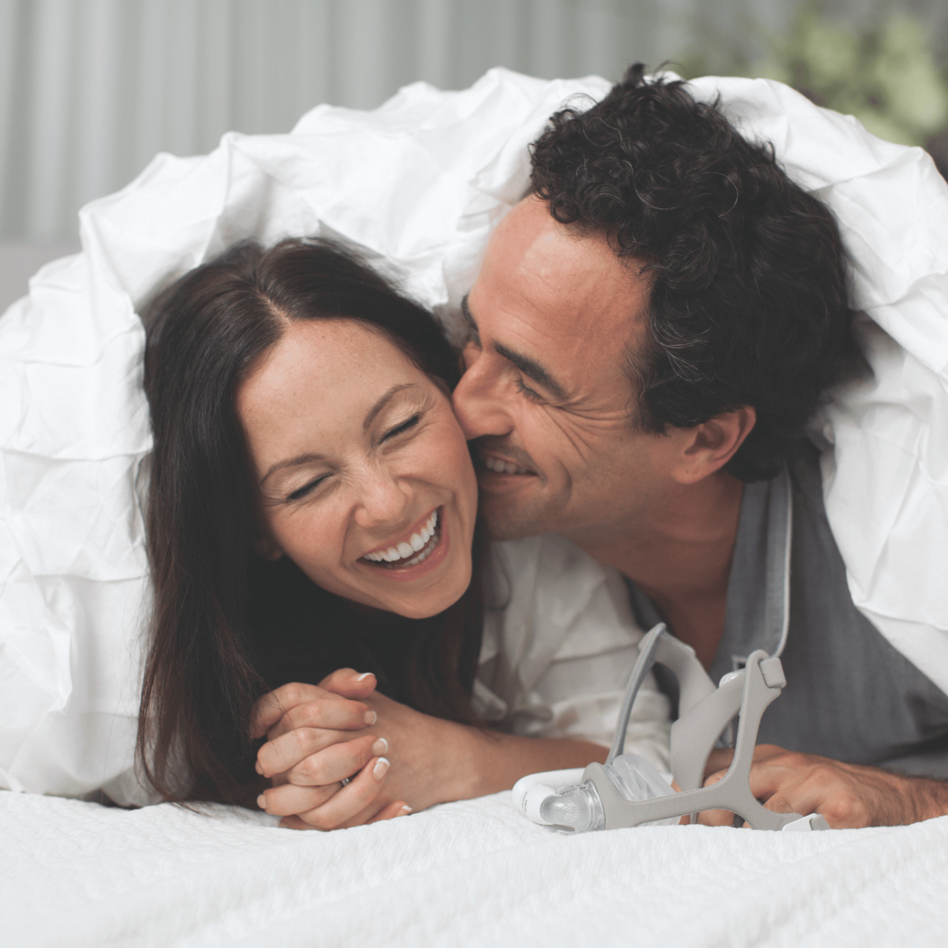 a couple laughs in bed under the covers with a wisp fabric nasal mask sitting on the bed in front of them